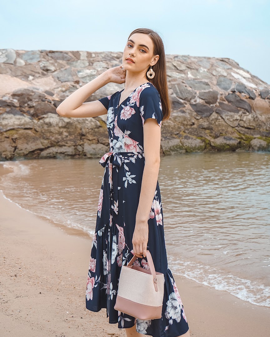 A model in a navy floral wrap dress and holding a handbag poses on a sandy beach.