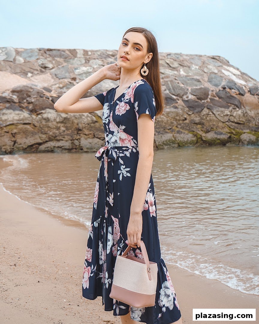 A model in a navy floral wrap dress and holding a handbag poses on a sandy beach.