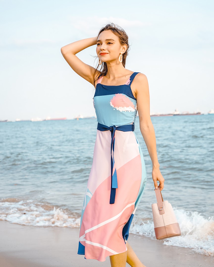 A woman in a colorful patterned dress and holding a bag stands by the sea.