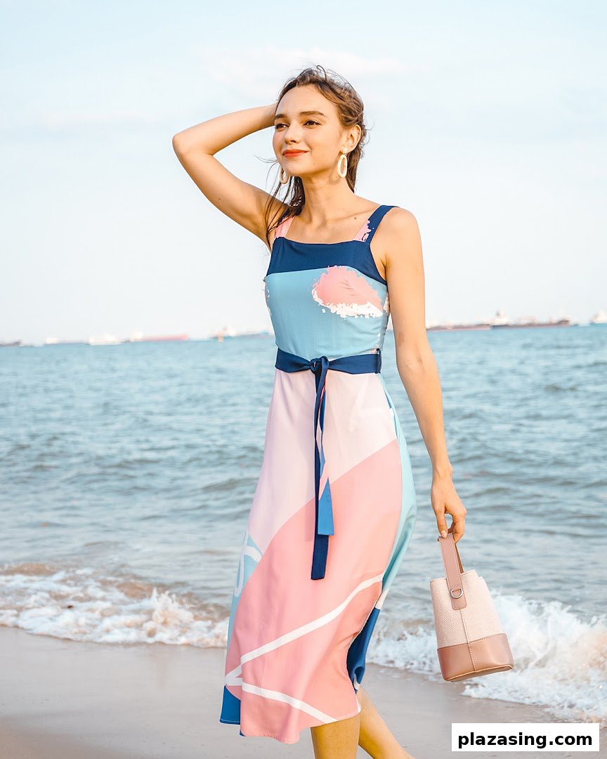 A woman in a colorful patterned dress and holding a bag stands by the sea.