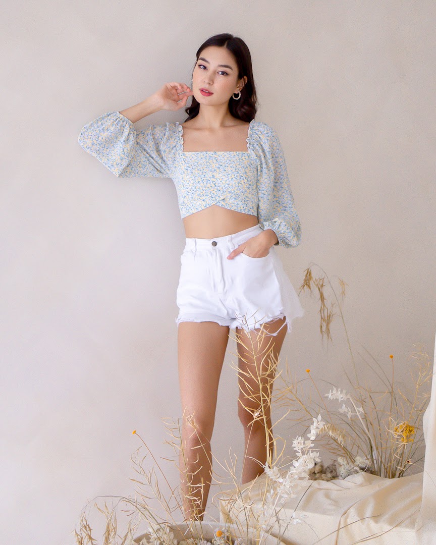A model showcases a light blue floral crop top and white shorts in a stylish, minimalist setting.