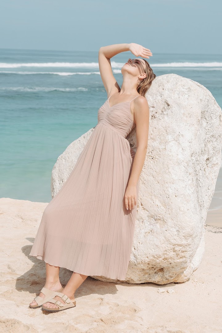 A woman in a flowing beige dress poses by a rock on a sunny beach with blue ocean.