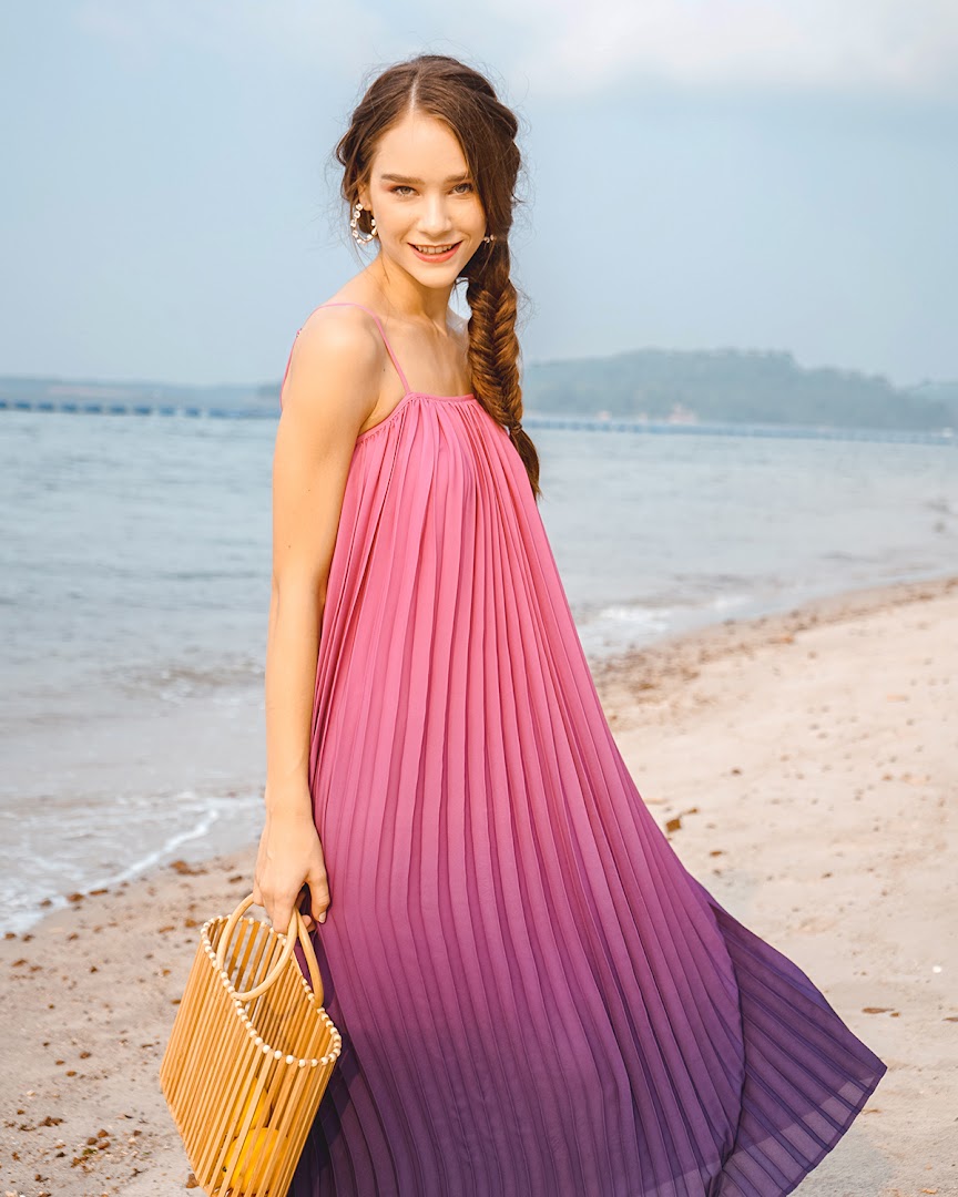 A woman in a pink and purple ombre pleated dress poses on a beach with a wooden bag.
