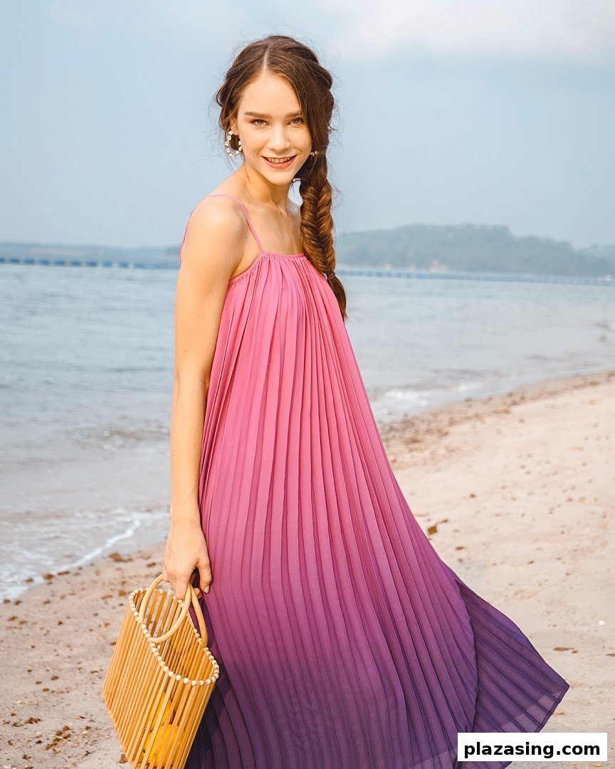 A woman in a pink and purple ombre pleated dress poses on a beach with a wooden bag.