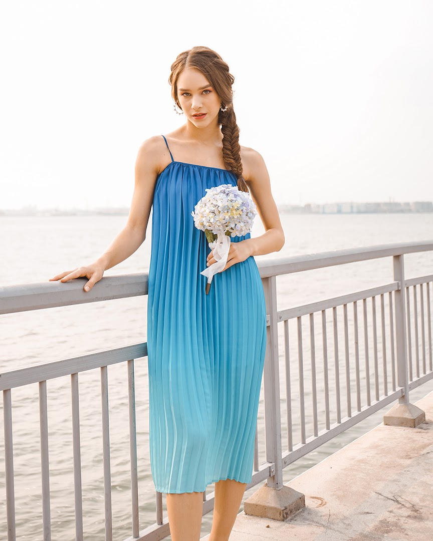 A woman in a blue ombre pleated dress holding a bouquet by the water.