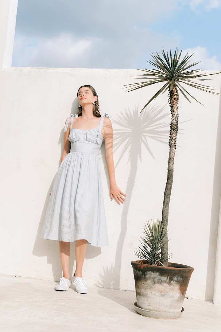 A woman in a light blue dress stands against a white wall with a potted plant.