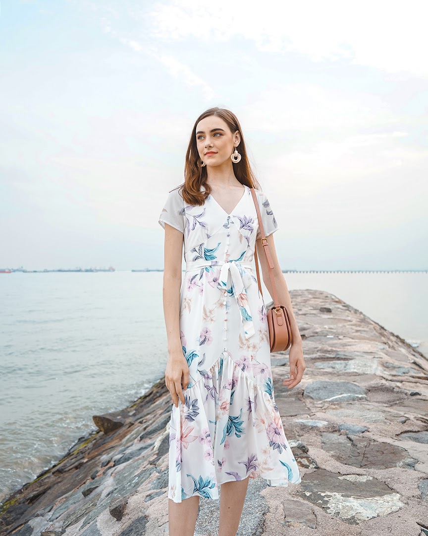 A woman in a white floral dress stands on a rocky pier by the sea.