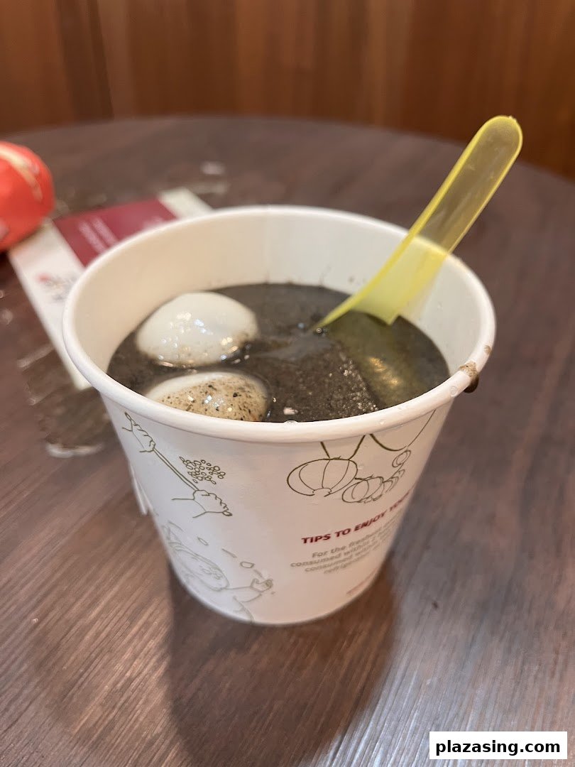 A cup of black sesame dessert with tangyuan and a yellow spoon on a wooden table.