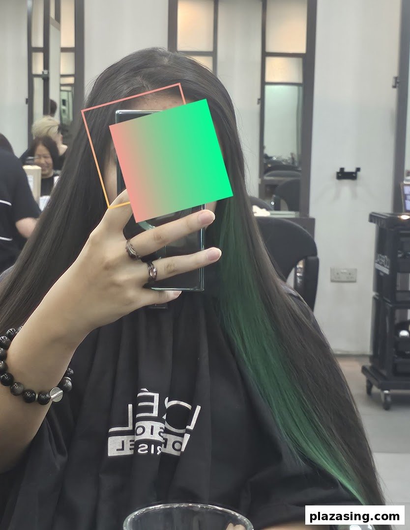 A person in a hair salon taking a selfie, showcasing long dark hair with vibrant green highlights.