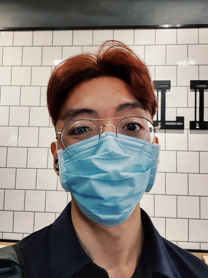 Close-up of a person with reddish hair, glasses, and a mask against a tiled wall.