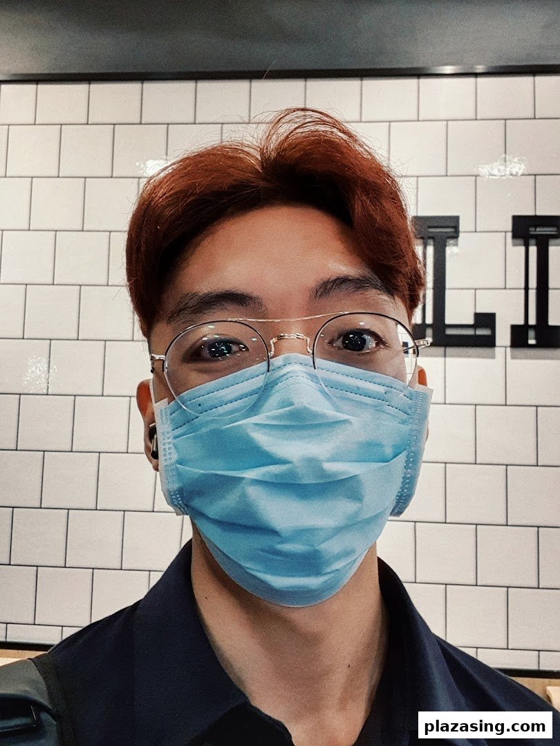 Close-up of a person with reddish hair, glasses, and a mask against a tiled wall.