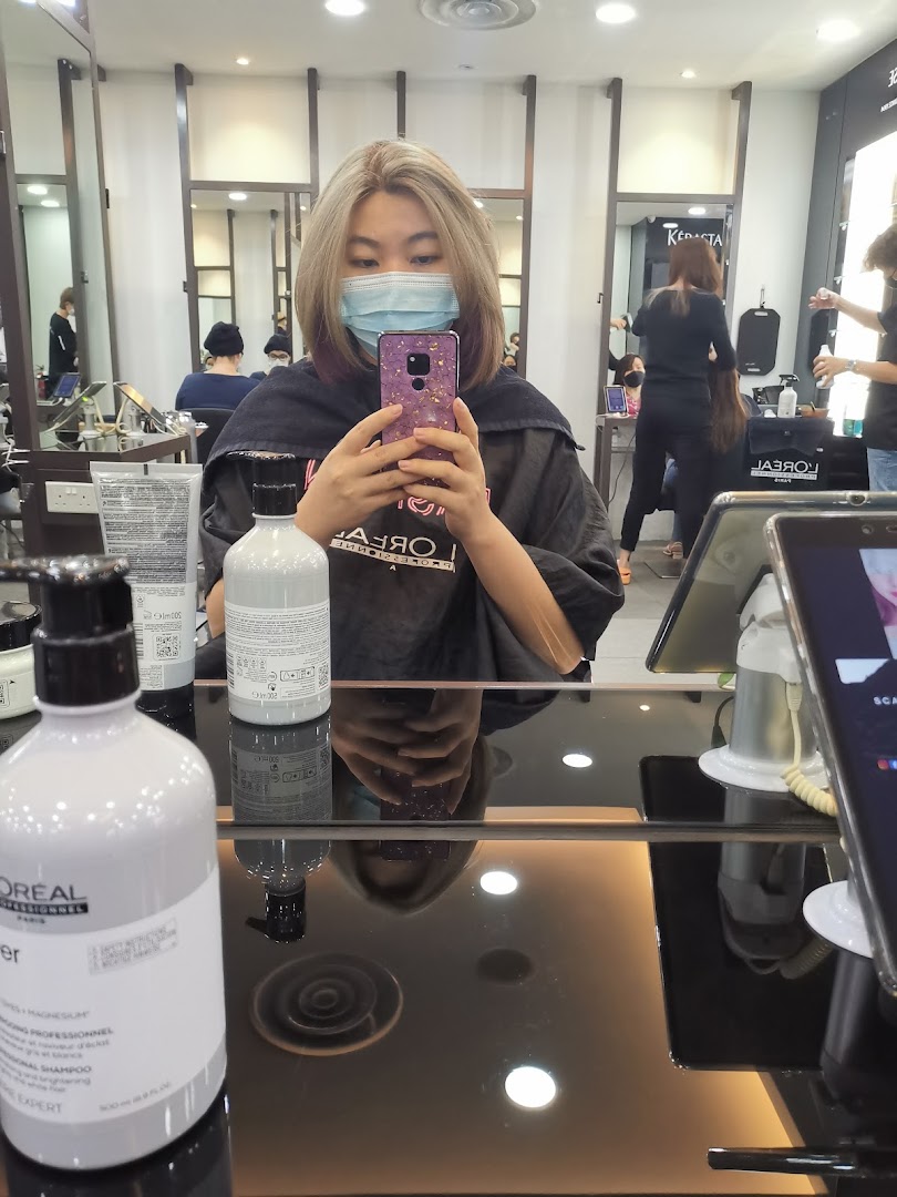A customer in a hair salon chair, wearing a mask, taking a selfie with hair products on the table.