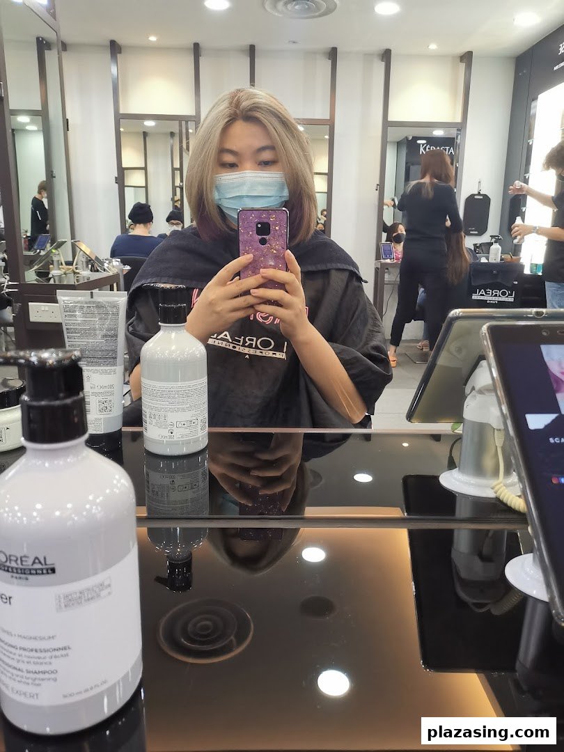 A customer in a hair salon chair, wearing a mask, taking a selfie with hair products on the table.