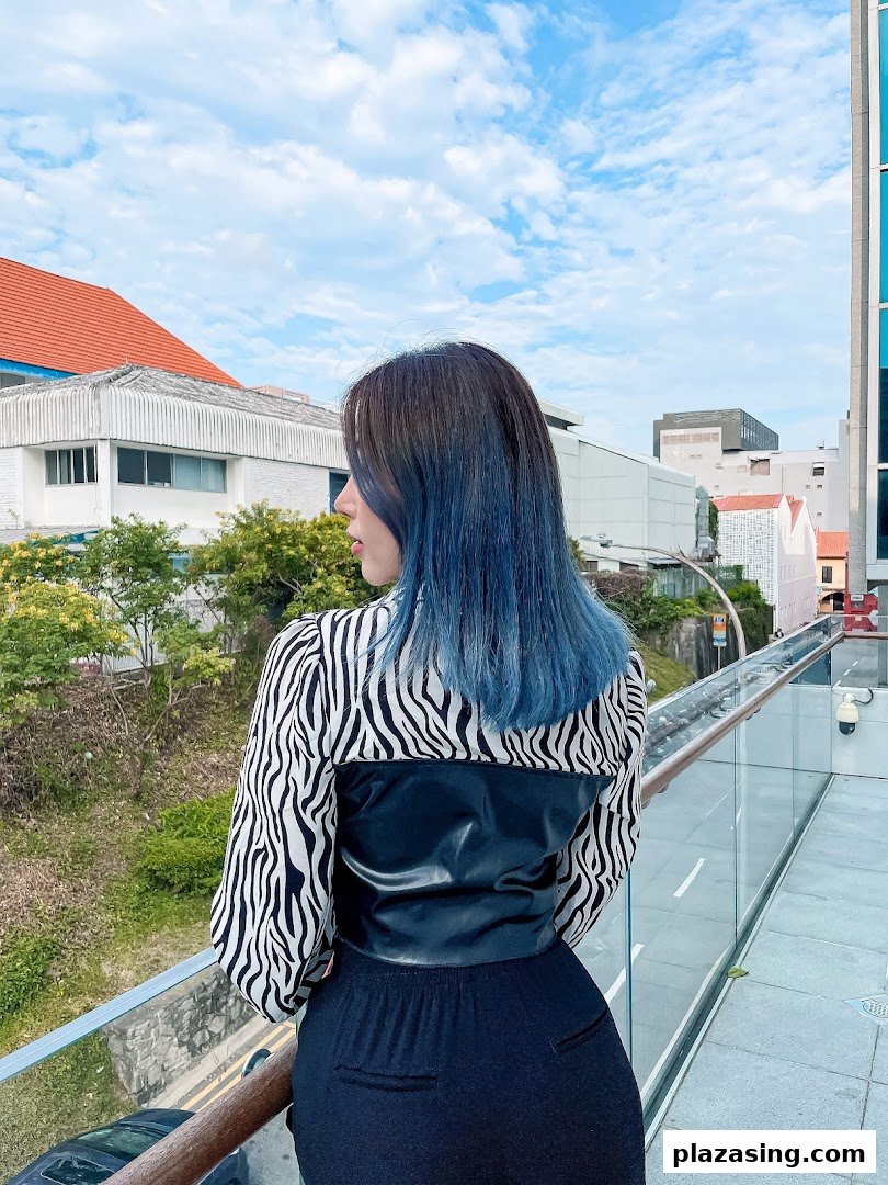 A person with vibrant blue hair stands on a bridge, looking out at the city skyline.