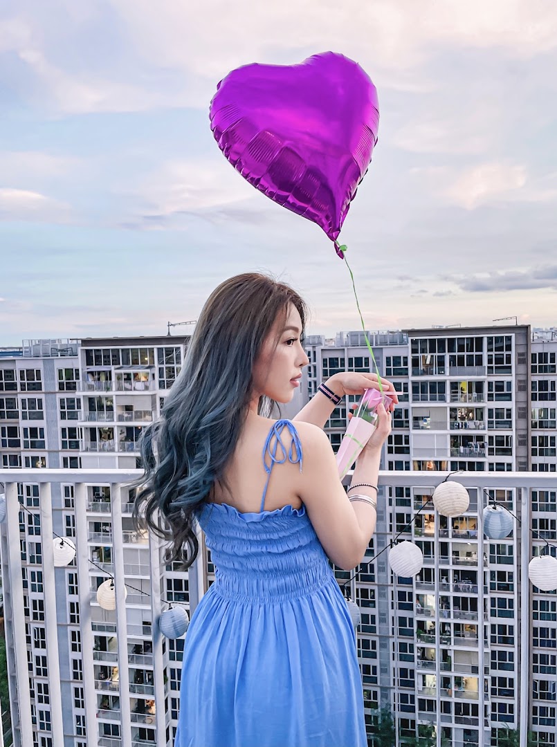 A woman with styled hair holding a purple heart balloon and a gift box on a balcony.