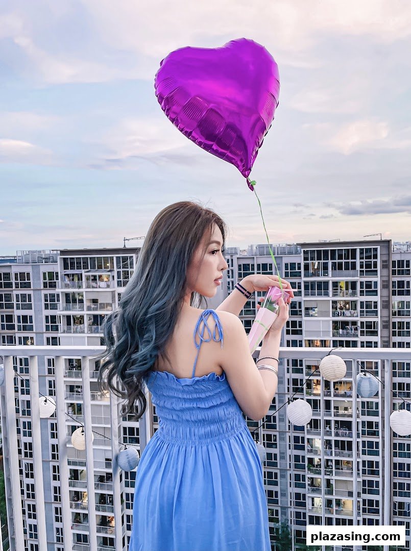 A woman with styled hair holding a purple heart balloon and a gift box on a balcony.