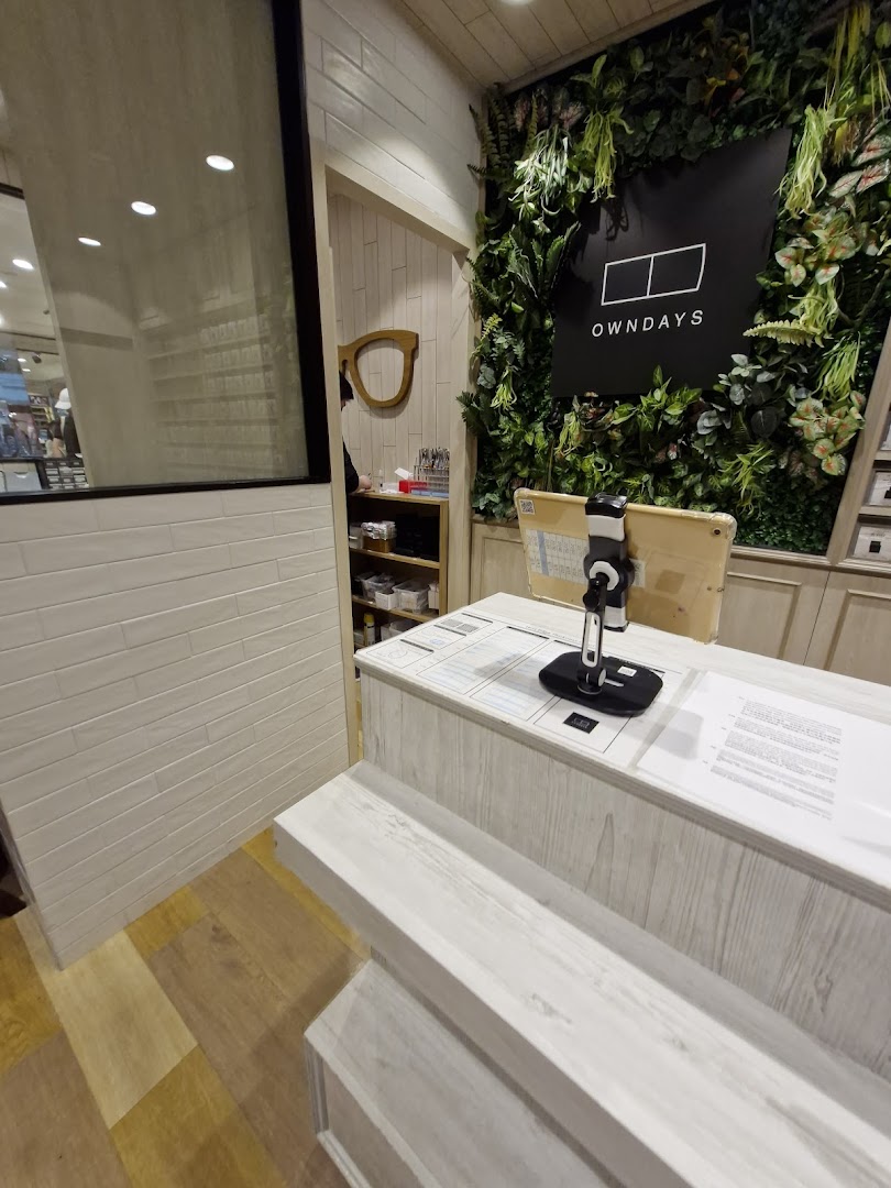 An interior view of OWNDAYS, featuring a modern counter and a lush green wall with the brand logo.