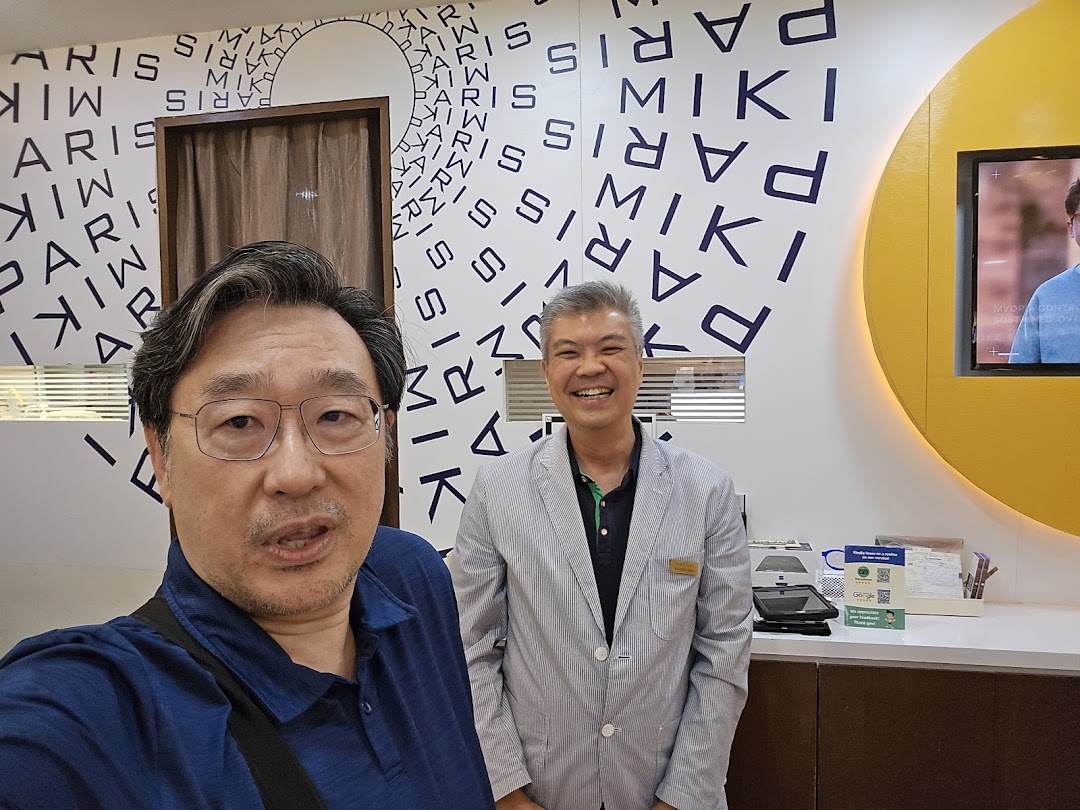Two men smiling inside Paris Miki optician shop, featuring branded wall decor and eye care displays.