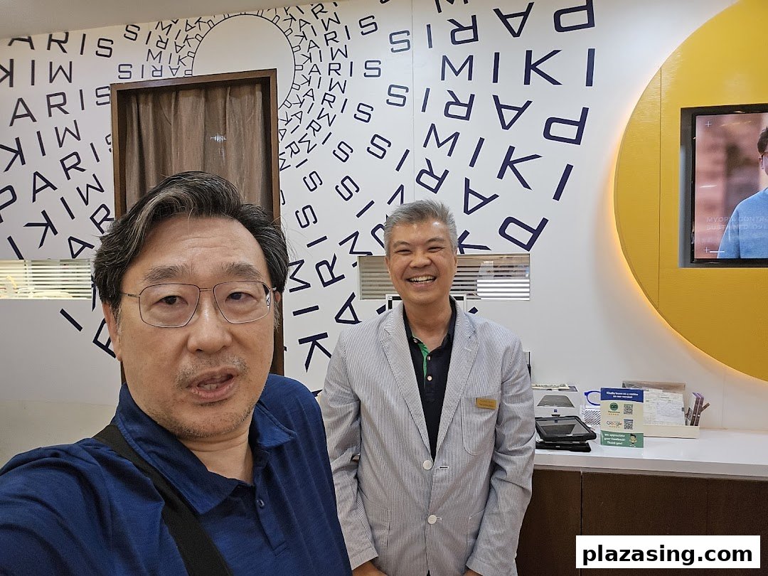 Two men smiling inside Paris Miki optician shop, featuring branded wall decor and eye care displays.