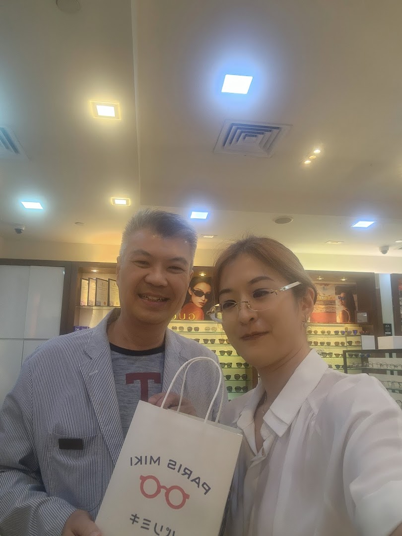 Two smiling individuals, one holding a Paris Miki bag, inside an optical shop with eyewear displays.