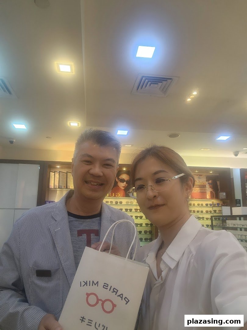 Two smiling individuals, one holding a Paris Miki bag, inside an optical shop with eyewear displays.