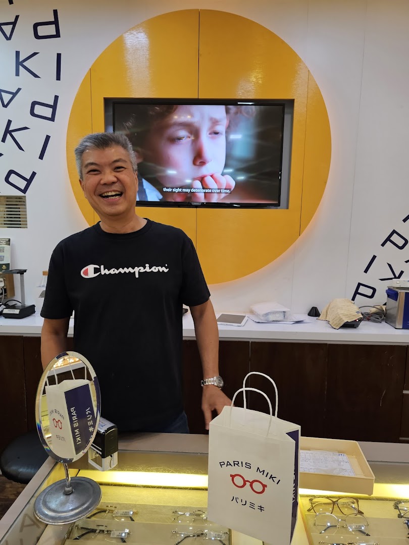 A smiling staff member stands behind a counter in an optician's shop, featuring eyewear displays.