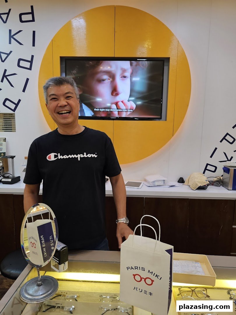 A smiling staff member stands behind a counter in an optician's shop, featuring eyewear displays.
