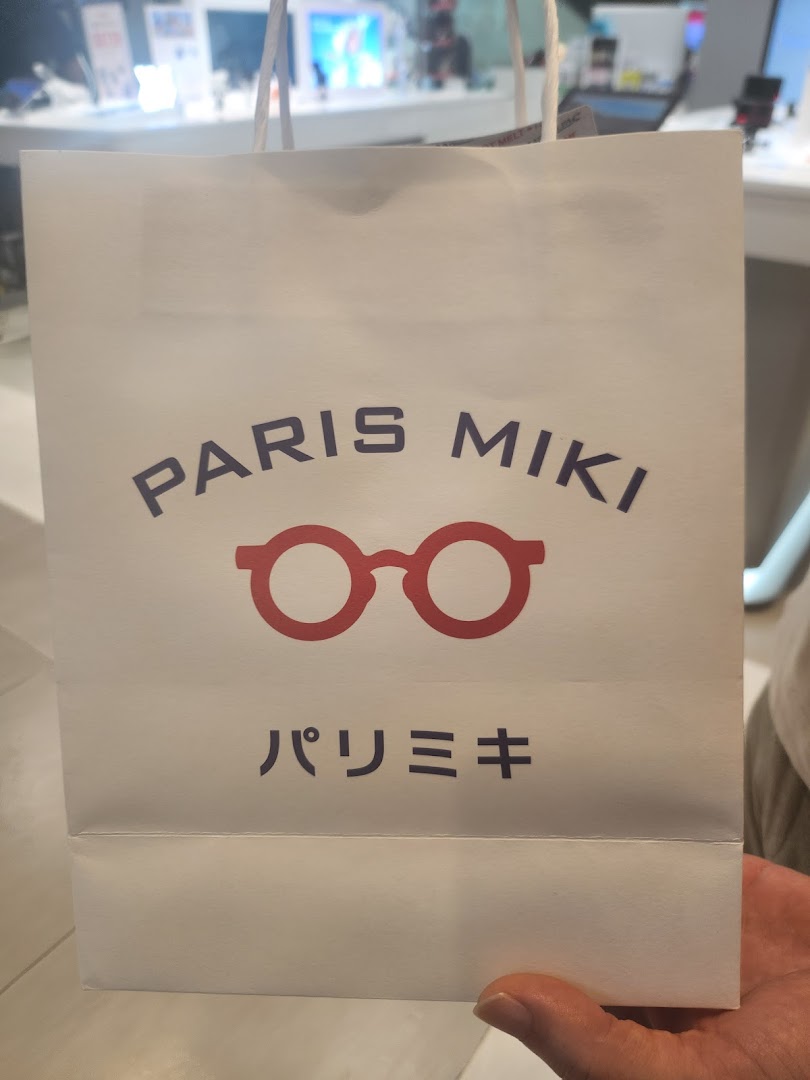 A white Paris Miki shopping bag featuring a red glasses logo, held in a retail environment.
