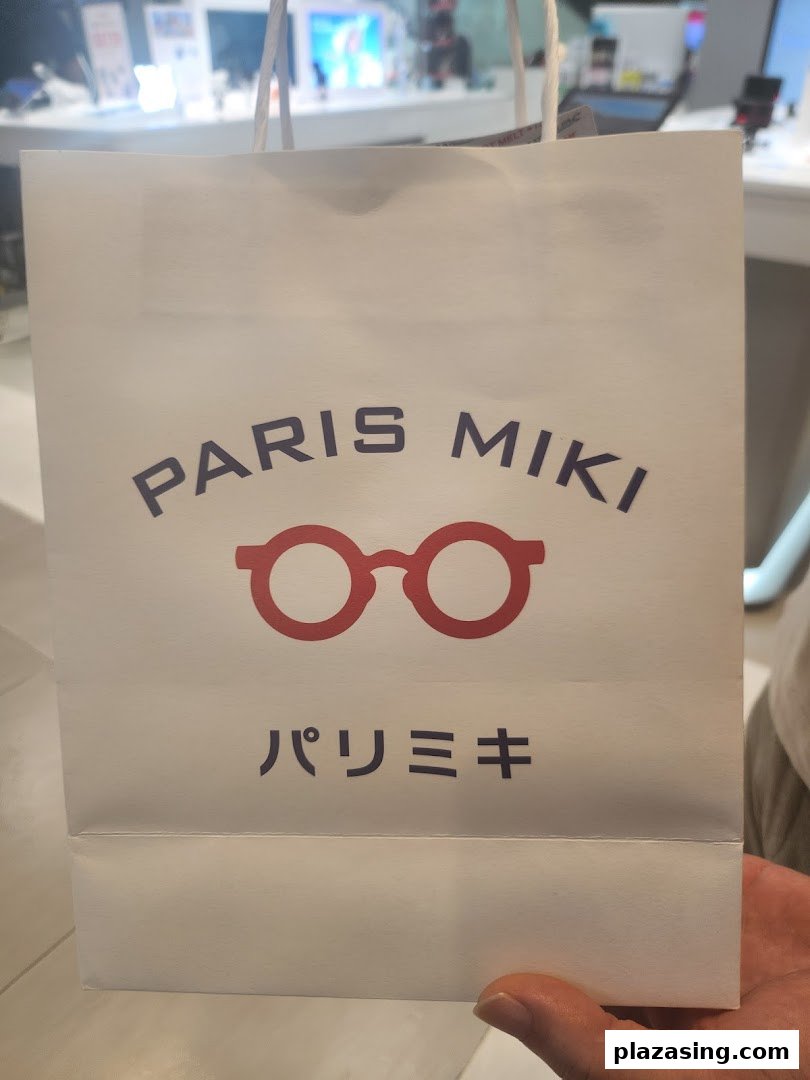 A white Paris Miki shopping bag featuring a red glasses logo, held in a retail environment.
