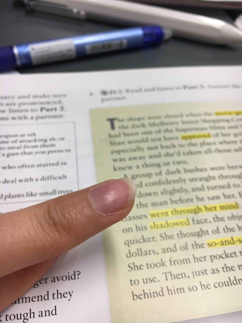 A close-up shot of a finger pointing at highlighted text in an open book, with a pen visible in the background.