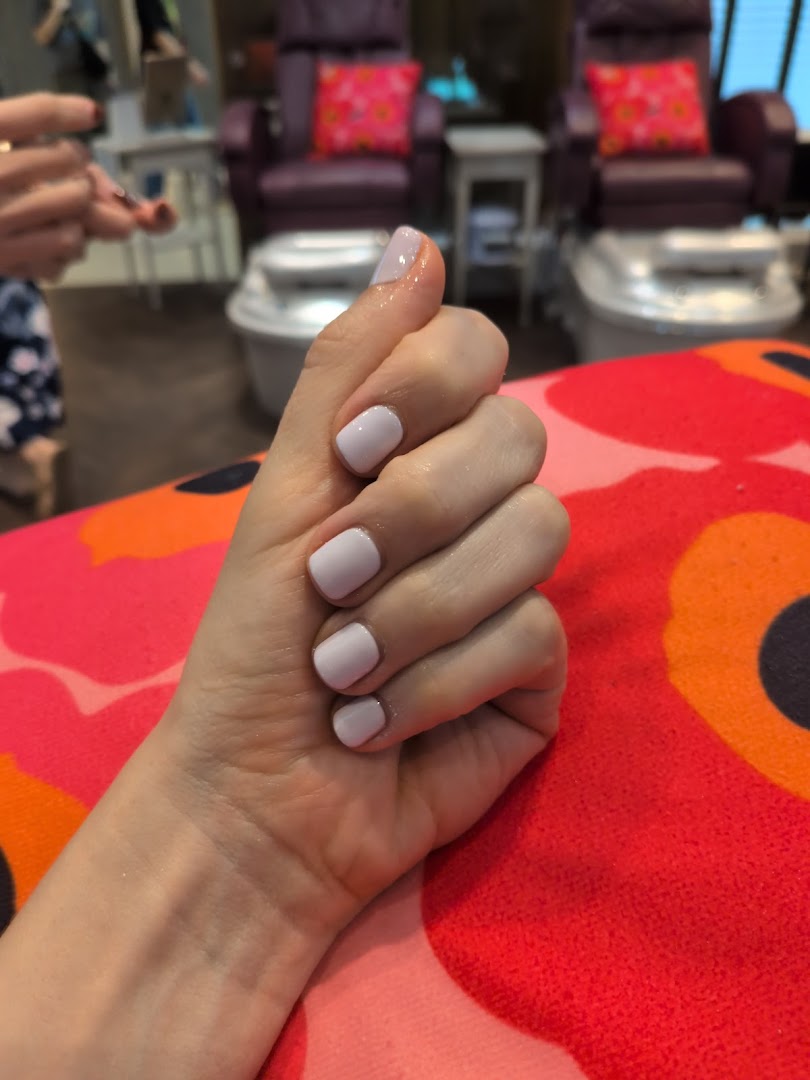 A hand with freshly painted light purple nails rests on a vibrant patterned cushion, with pedicure chairs in the background.