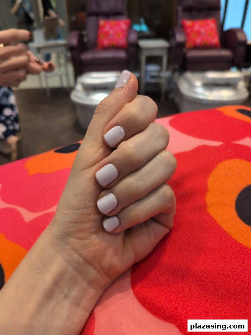 A hand with freshly painted light purple nails rests on a vibrant patterned cushion, with pedicure chairs in the background.