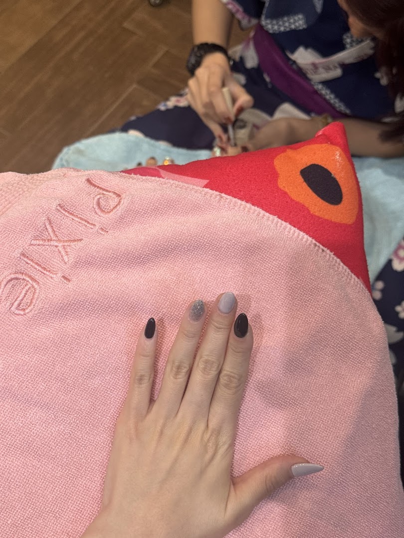 A customer's hand with freshly painted nails resting on a pink towel, with a technician performing a pedicure in the background.