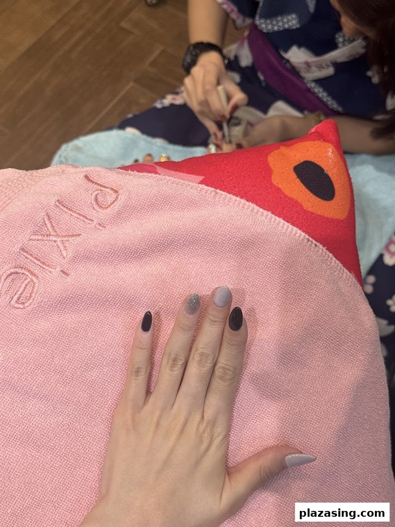 A customer's hand with freshly painted nails resting on a pink towel, with a technician performing a pedicure in the background.