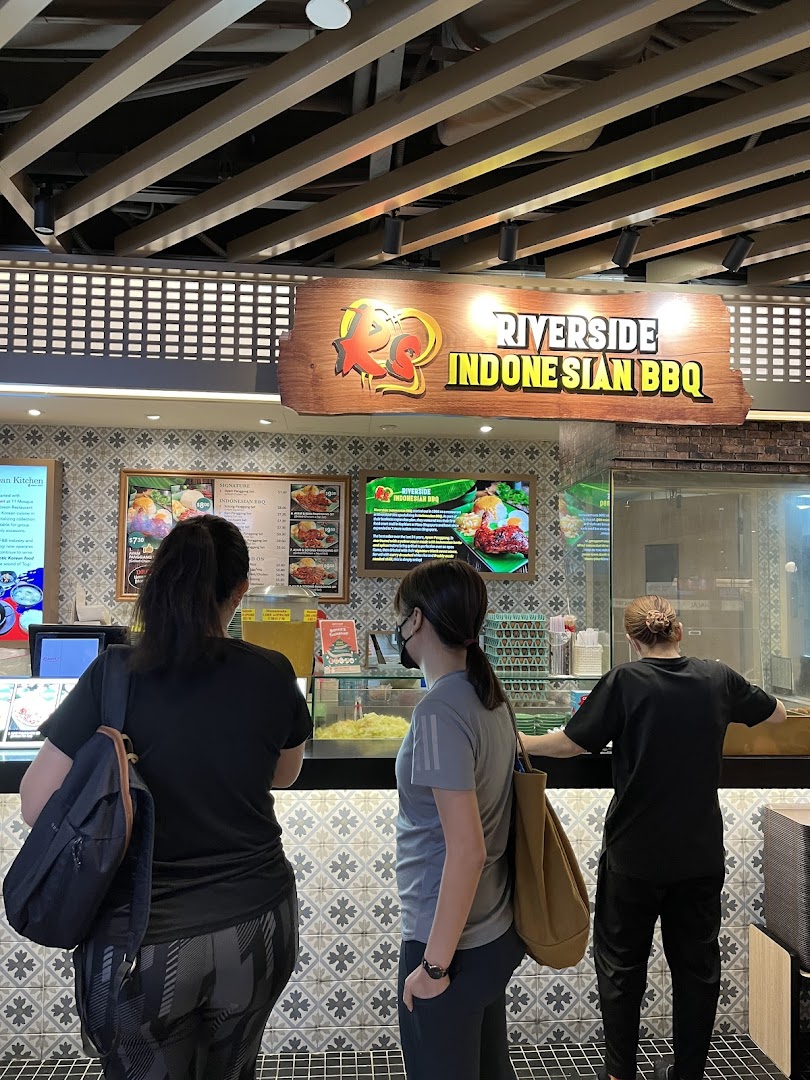 Customers queue at Riverside Indonesian BBQ counter, featuring vibrant menu displays and a welcoming atmosphere.