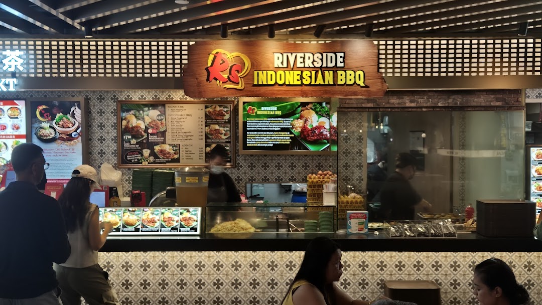 A bustling food stall, Riverside Indonesian BBQ, with customers, staff, and vibrant menu displays.