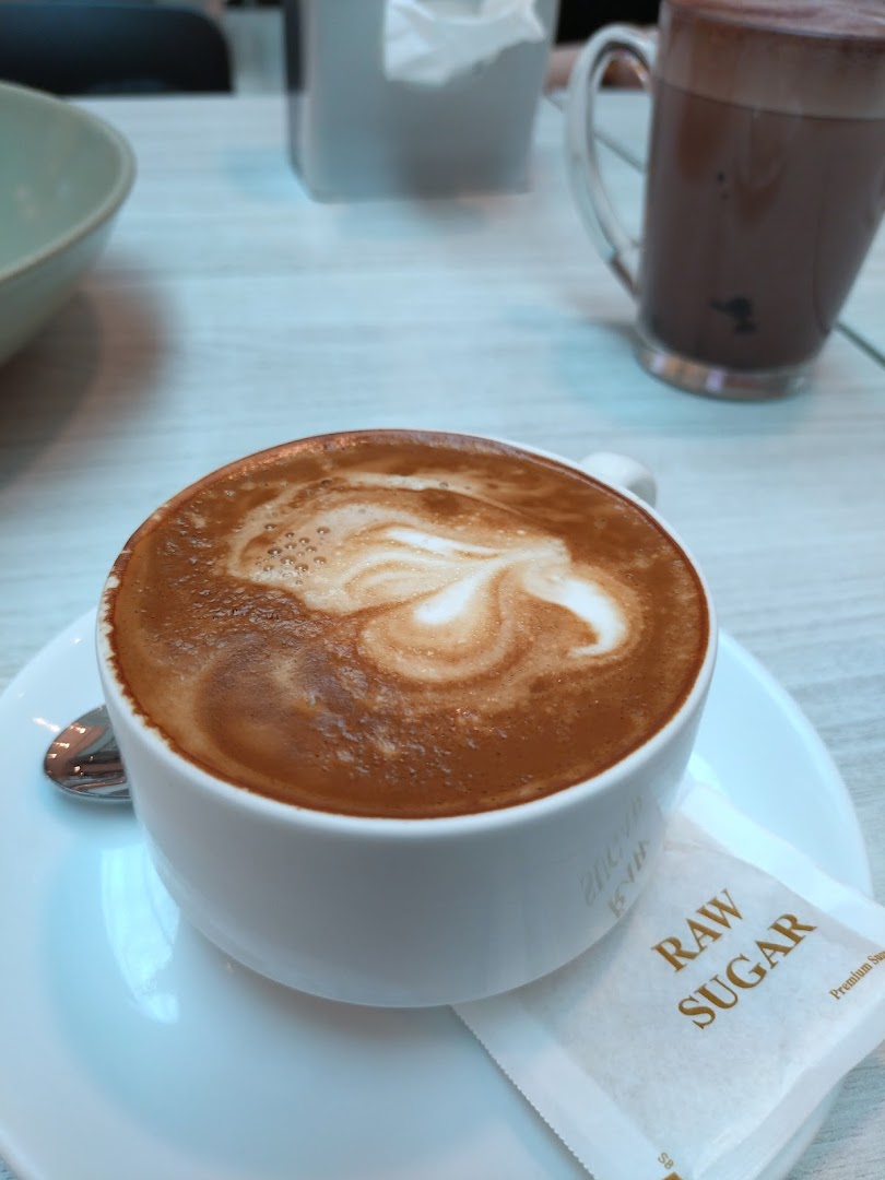 A close-up of a latte with latte art, a raw sugar packet, and another drink in the background.