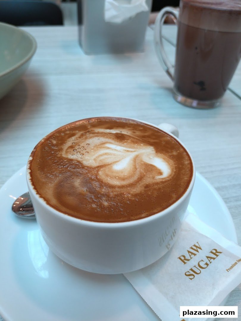A close-up of a latte with latte art, a raw sugar packet, and another drink in the background.