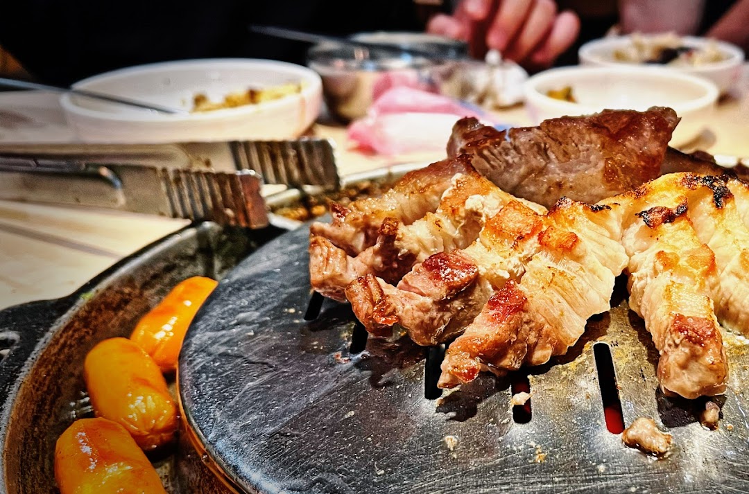 Close-up of sizzling grilled pork belly and sausages on a hot plate at a Korean BBQ restaurant.