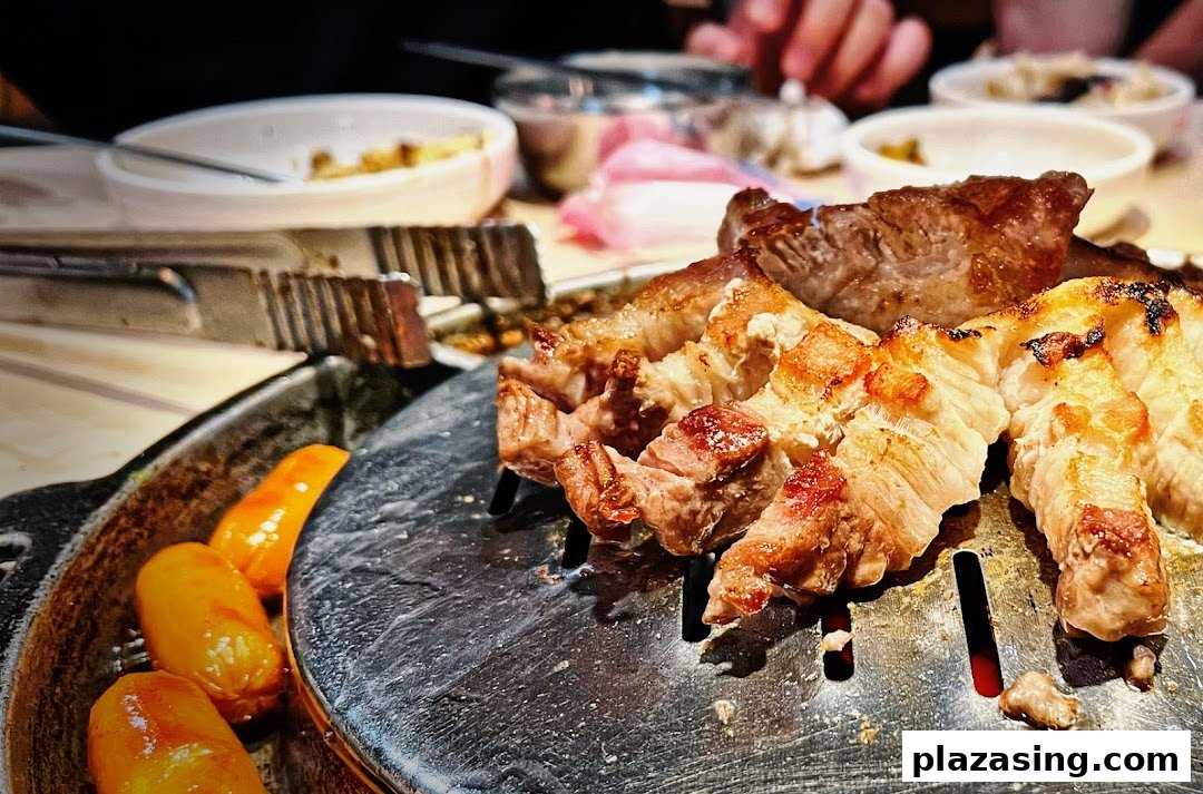 Close-up of sizzling grilled pork belly and sausages on a hot plate at a Korean BBQ restaurant.