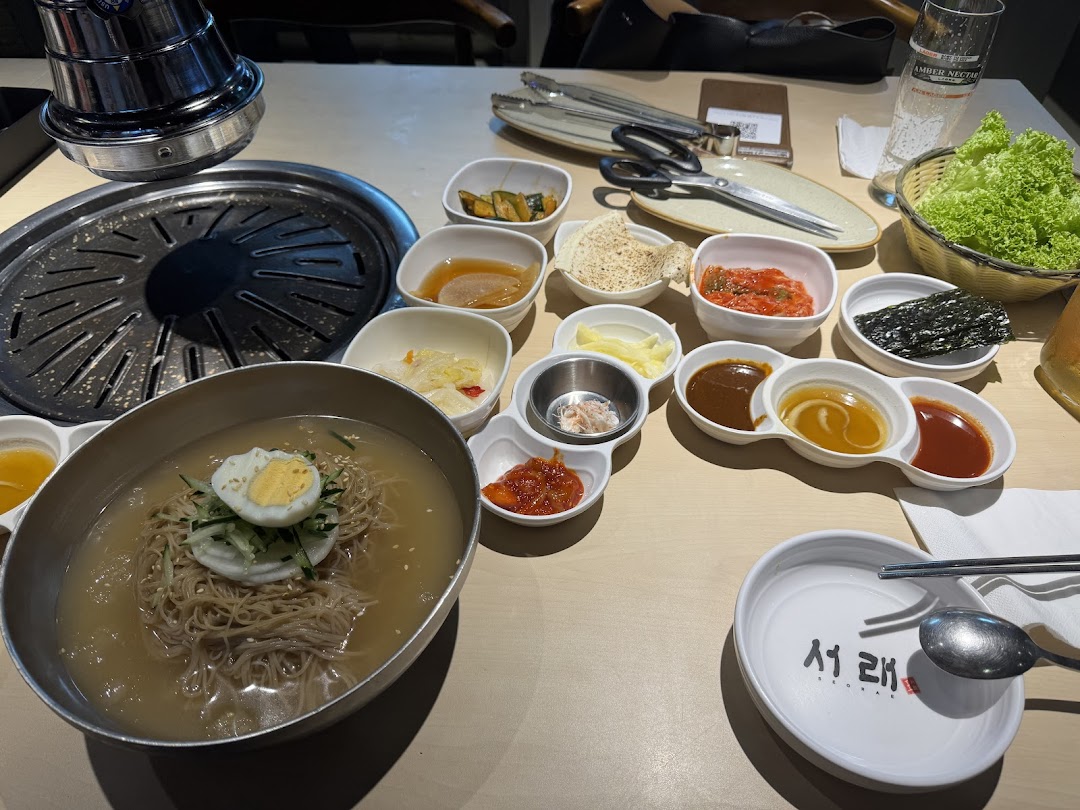 A table laden with a variety of Korean dishes, including cold noodles, banchan, and a grill.