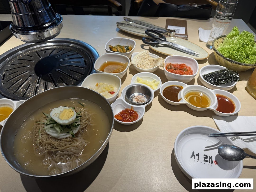 A table laden with a variety of Korean dishes, including cold noodles, banchan, and a grill.