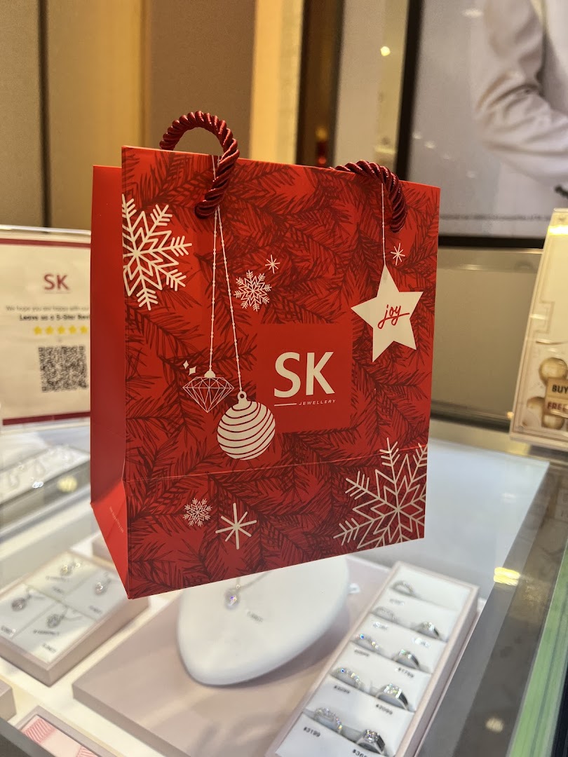 A festive red SK Jewellery shopping bag displayed amidst sparkling jewellery in a retail setting.