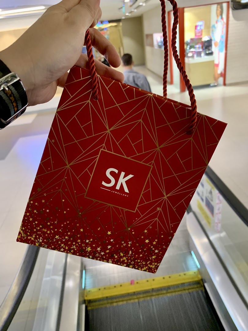 A person holds a festive red SK Jewellery shopping bag with gold patterns, near an escalator.