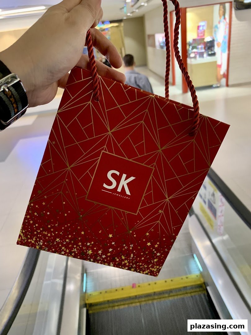 A person holds a festive red SK Jewellery shopping bag with gold patterns, near an escalator.