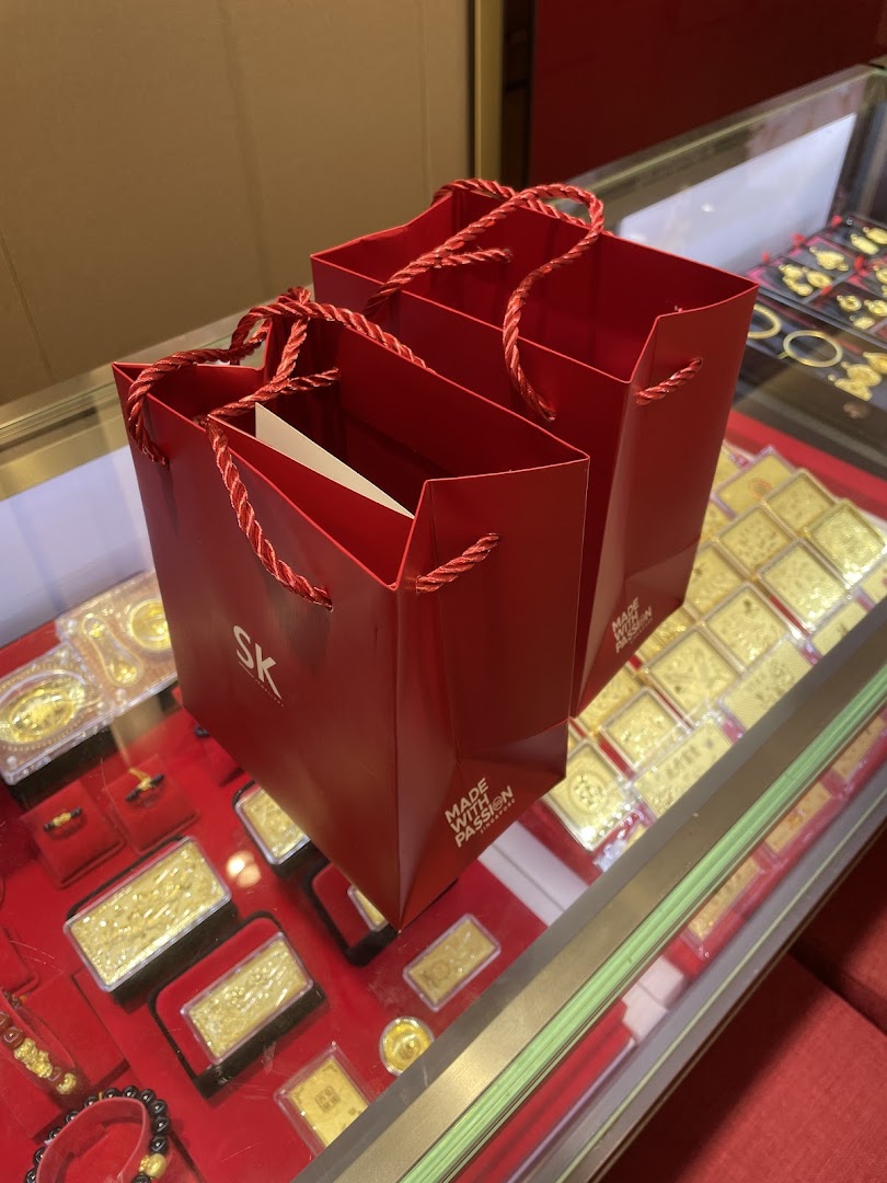Two red SK Jewellery shopping bags placed on a display case filled with various gold items.