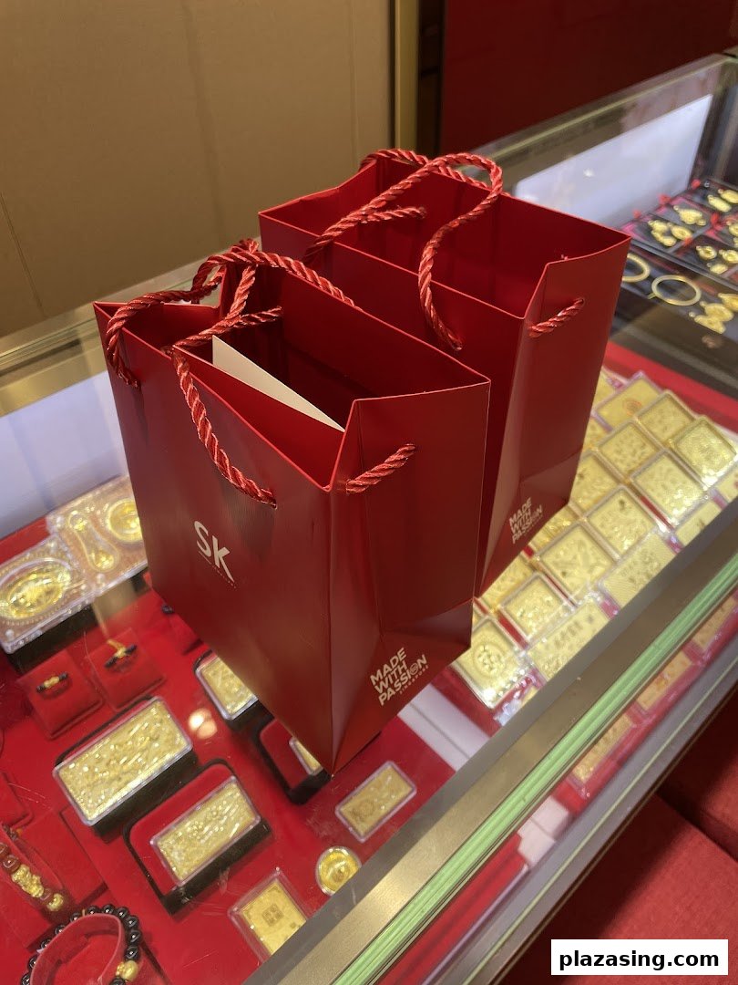 Two red SK Jewellery shopping bags placed on a display case filled with various gold items.