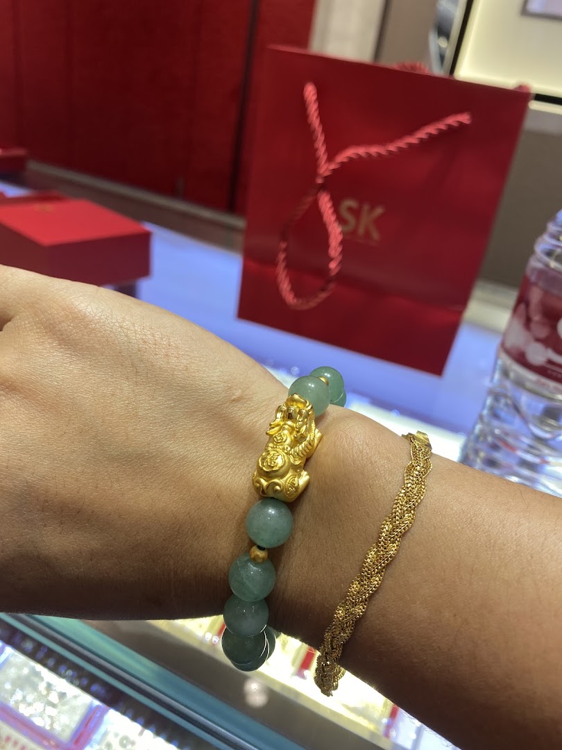 A person's arm showcasing a jade and gold Pixiu bracelet alongside a braided gold bracelet, with an SK Jewellery bag in the background.