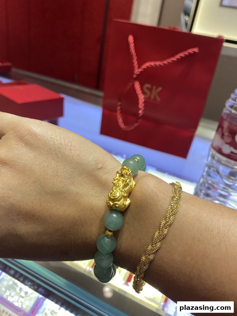 A person's arm showcasing a jade and gold Pixiu bracelet alongside a braided gold bracelet, with an SK Jewellery bag in the background.