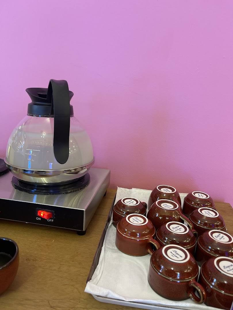 A kettle on a hot plate and a tray of ceramic cups against a vibrant pink wall, offering refreshments.